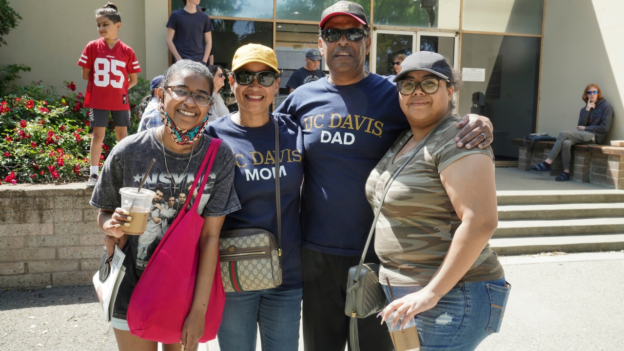 Four people stood in front of a building. The woman second from the left is wearing a UC Davis Mom T-Shirt, and the man to the right of her is wearing a UC Davis Dad T-Shirt.