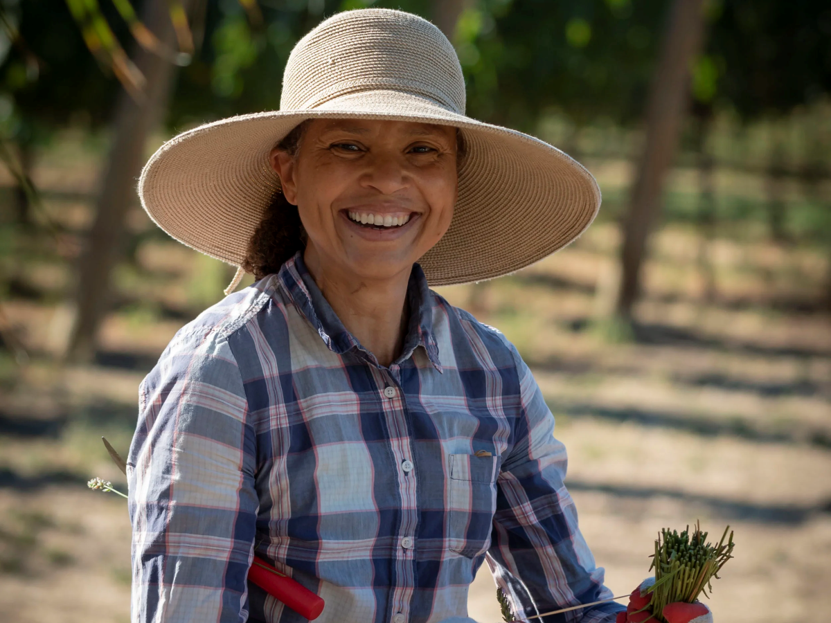 Smiling woman in wide-brim hat and plaid shirt holding freshly picked carrots in a sunny field