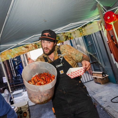 Man in cap and apron carrying a large bowl of crawfish under an outdoor tent