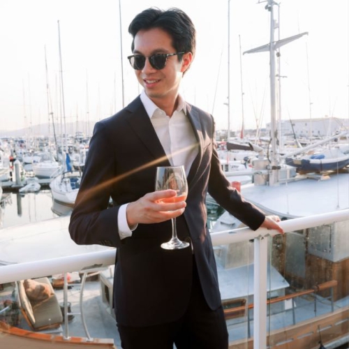 Man in suit and sunglasses holding a wine glass on a marina deck at sunset