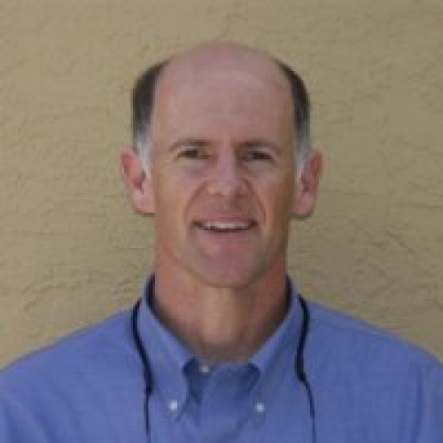 Headshot of a smiling middle-aged balding man in blue shirt against tan wall