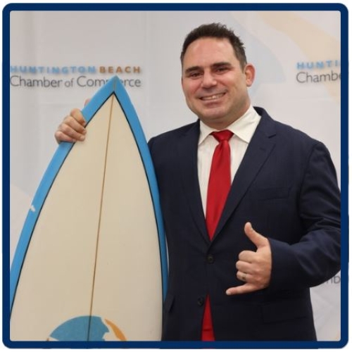 Smiling man in dark suit and red tie holding a surfboard and making a shaka gesture