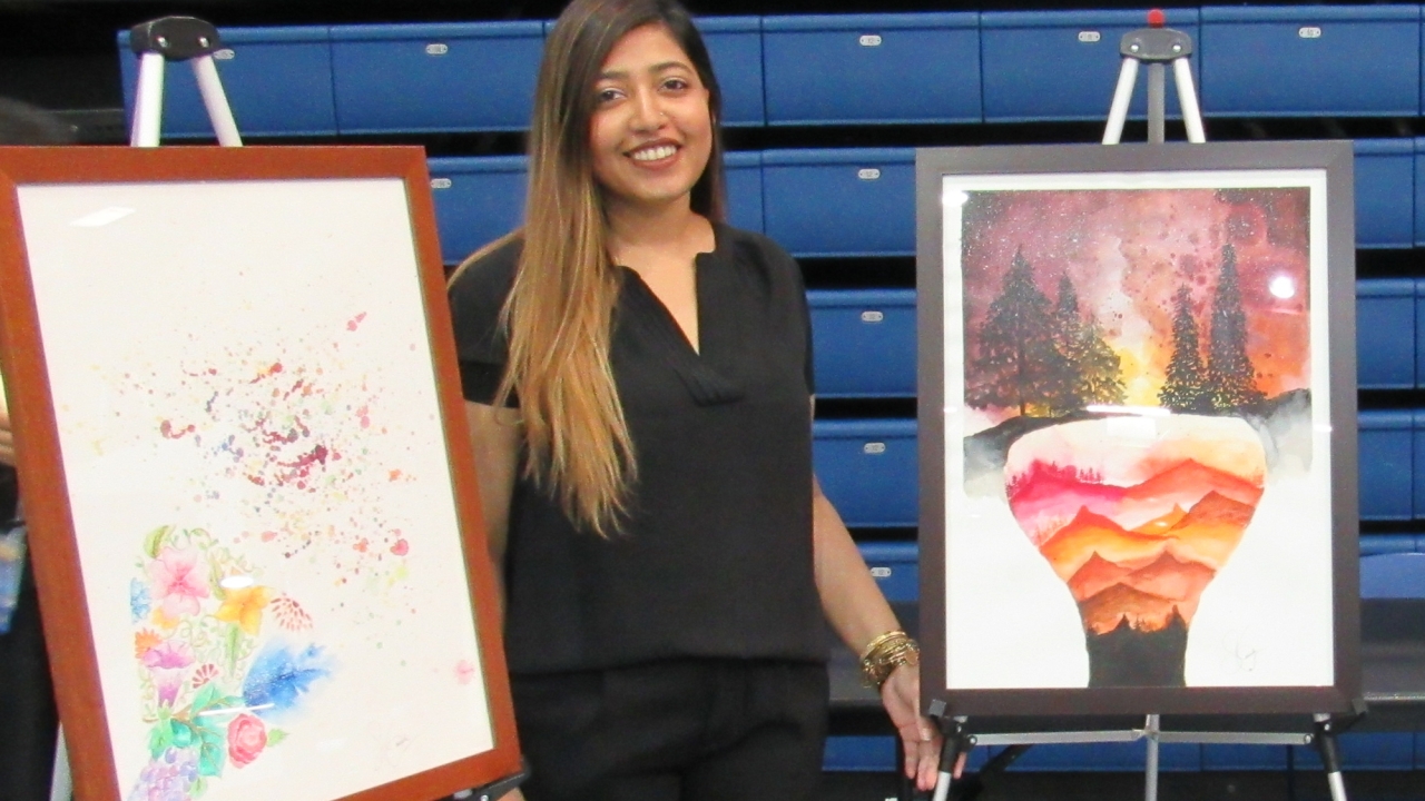 A woman with long brown hair, wearing a black outfit, stands smiling in front of three framed paintings displayed on easels in an indoor exhibition space. The artwork on the left features an abstract composition with splashes of color and floral elements. The middle piece portrays a surreal landscape with layered, colorful hills inside a floating shape against a night sky with trees. The artwork on the right consists of a dark blue abstract form with splattered paint on a white background. The setting inclu