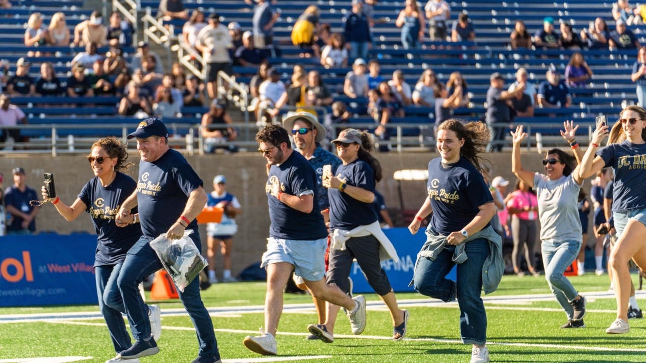 In the forefront, 8 people are in the motion of running on the football field in Aggie Pride blue shirts. In the background are people standing on the sidelines of the field and you can also see fans sitting the stands.