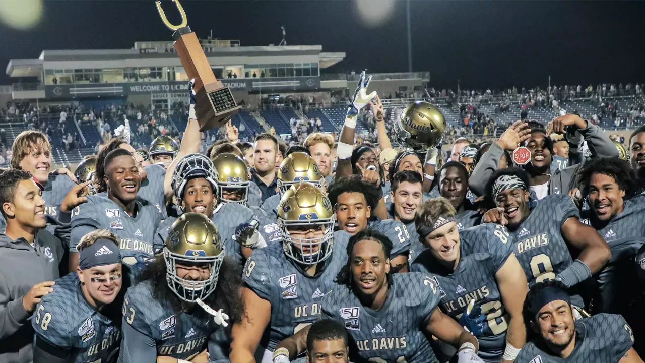 UC Davis football players wearing grey uniforms holding up the Golden Horseshoe Trophy in celebration