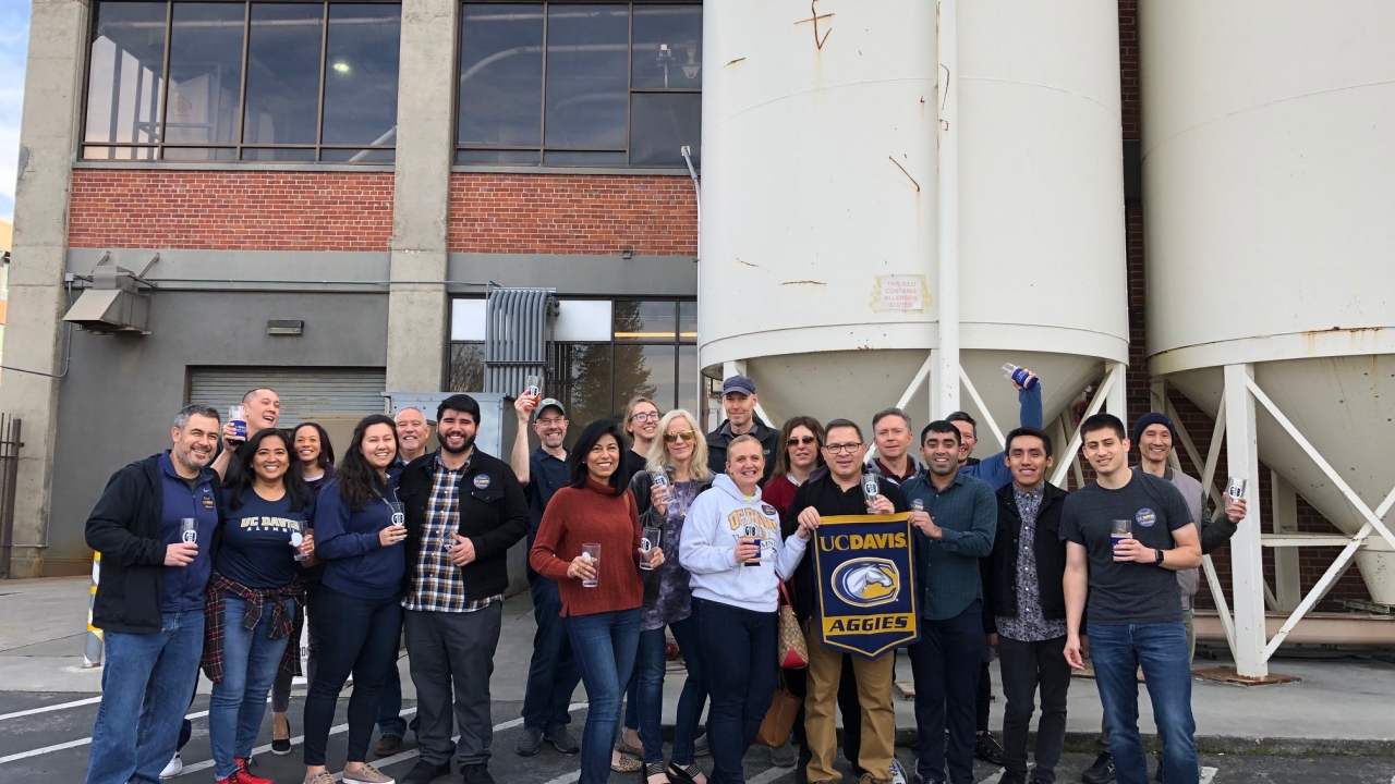 Tour group of 21 people standing outside a factory cheerings to the camera with beer glasses, with two people holding a UC Davis Aggies banner