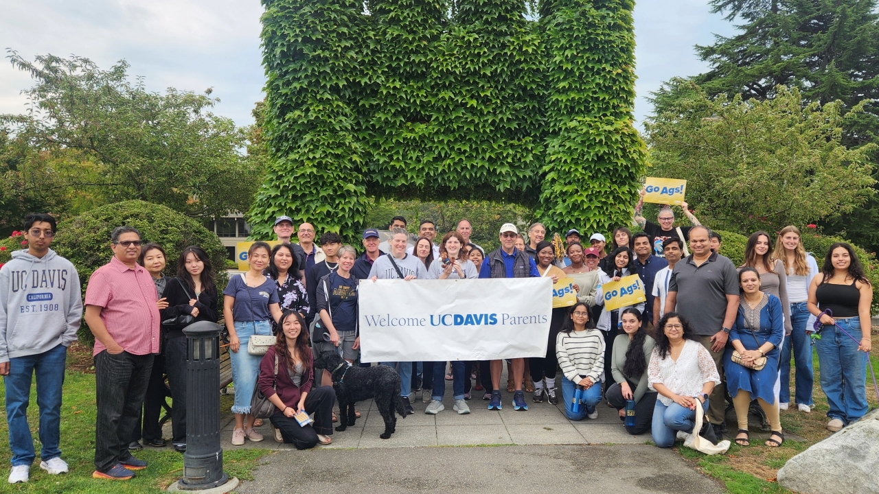 Diverse group smiling outdoors, holding a wide banner in front of tall green foliage