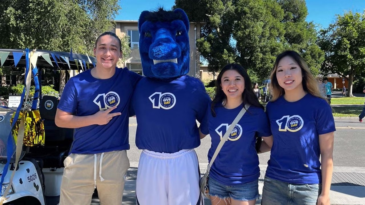 Four smiling people outside — three women in blue "10" shirts and a tall blue mascot