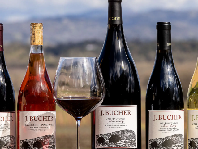 J. Bucher wine bottles and a glass of red wine on a wooden table with rolling hills background