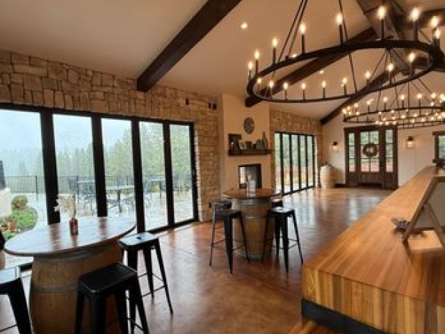 Warm-lit winery tasting room with wooden bar, barrel-top tables, chandeliers, vineyard view.