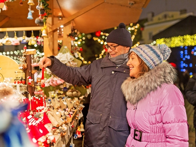 A couple smiles and points at decorations in a festive market adorned with lights.