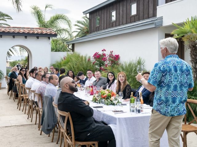 Outdoor courtyard dinner with guests at a long table, man in blue giving a toast