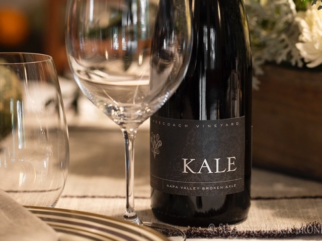 Wine bottle labeled "KALE" beside empty wine glass on a warmly lit table with flowers