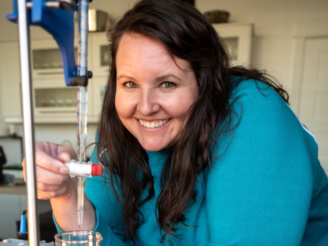 Smiling woman in teal sweater leaning over lab bench with pipette and glassware