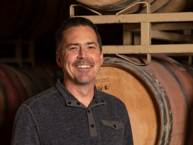 Smiling man in dark sweater standing before stacked oak wine barrels