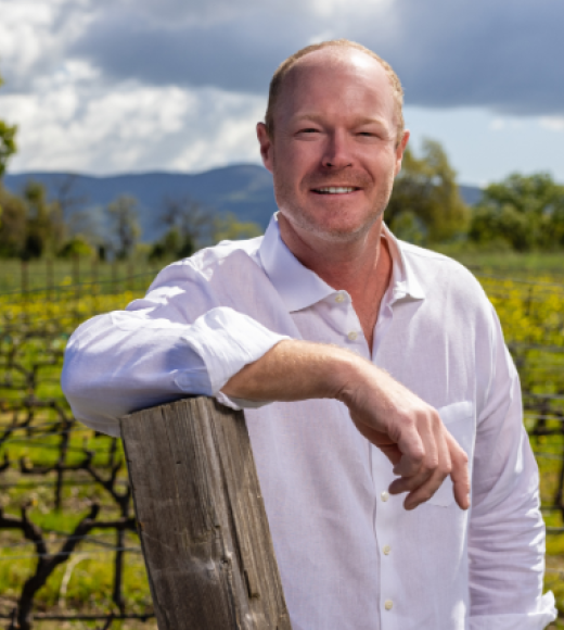 Smiling man in white shirt leaning on fence post in vineyard under cloudy sky, photo
