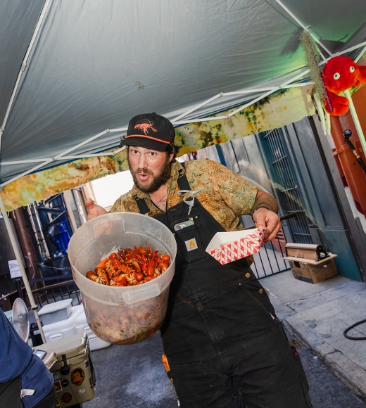 Man in cap and apron carrying a large bowl of crawfish under an outdoor tent