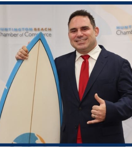 Smiling man in dark suit and red tie holding a surfboard and making a shaka gesture