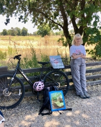 Margaret Ann Eldred leaning against a fence outdoors next to a road bicycle and a pink helmet with two paintings resting against the bicycle.