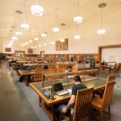 Photo of the main study room in the library, with stakes of books on the left and tables with students studying in the middle