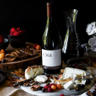 Still-life photograph of wine bottles, cheeses, grapes, figs and nuts on a dark table
