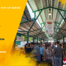 Crowd at a farmer's market under a roof with vendor stalls and colorful signage.
