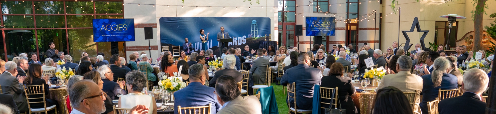 A group of guests dressed in elegant evening wear seated outside on Moss Patio around a nicely decorated round dinner table complete with place settings while facing the stage for the awards ceremony.