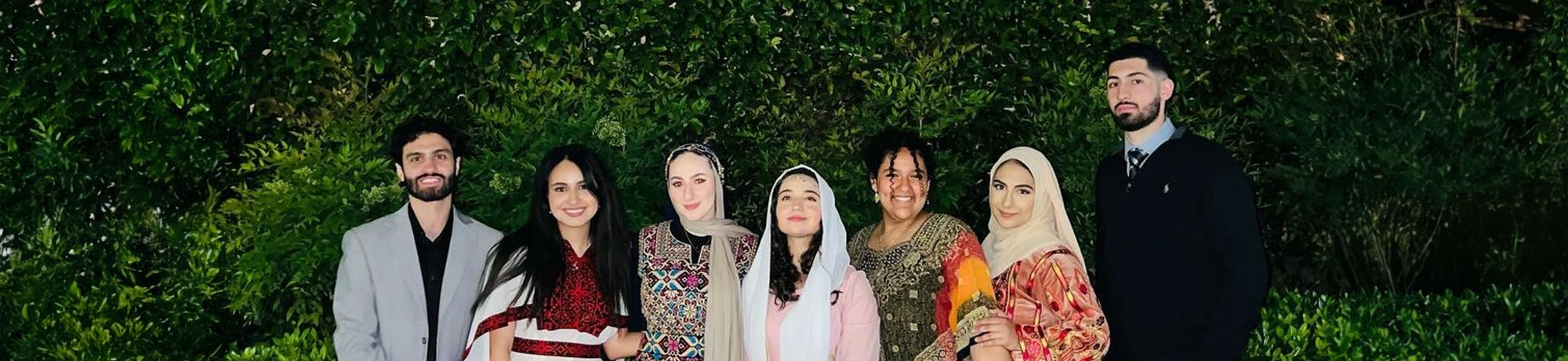 A group of students in the Arab Student Union at UC Davis pose in formal attire outdoors in front of greenery for an event.